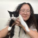Smiling person with glasses holding a black-and-white cat against a white background.