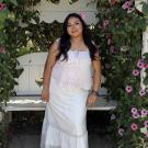 Smiling woman in white top and skirt standing by white bench beneath leafy arch of purple flowers