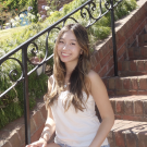 Smiling young woman with long hair sitting on sunlit brick steps by a wrought-iron railing