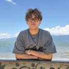 Young man wearing glasses and gray tee leaning on seaside wall, mountains and blue sky (photo)
