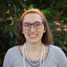 Smiling woman with glasses and orange headband, wearing pearls, garden background